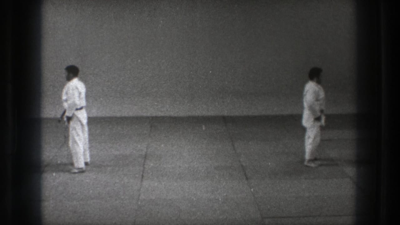 Two adult martial artists stand apart on a mat in Tokyo, 1971, seen in a grainy black-and-white 8mm frame.