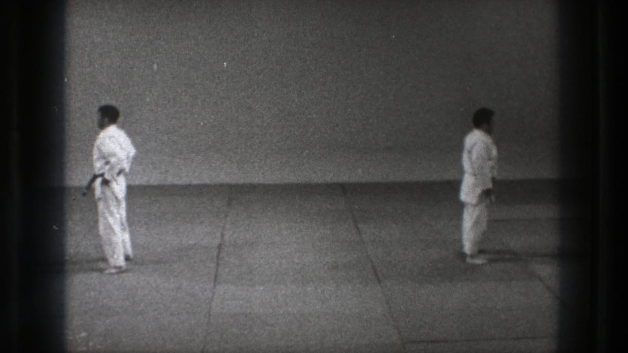 Two men in white gi stand apart on a plain indoor mat.