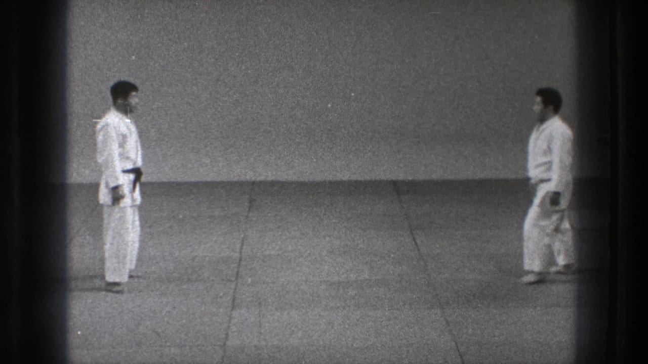 In the stated 1971 Tokyo setting, two adult martial-arts practitioners in white gi face one another across a plain matted training area.