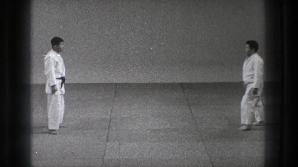 In the stated 1971 Tokyo setting, two adult martial-arts practitioners in white gi face one another across a plain matted training area.