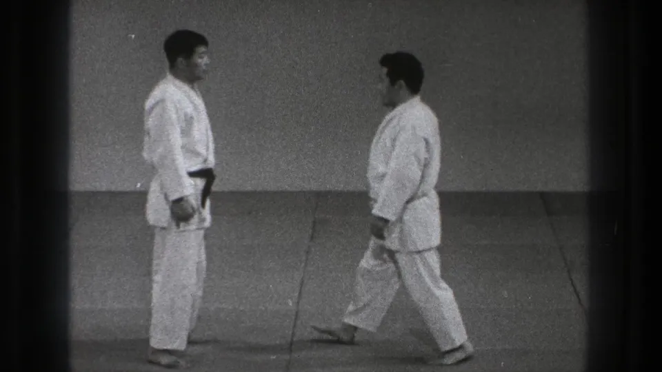 Close view of two adult practitioners in white gi on a mat, with one stepping forward as the paired technique setup begins.