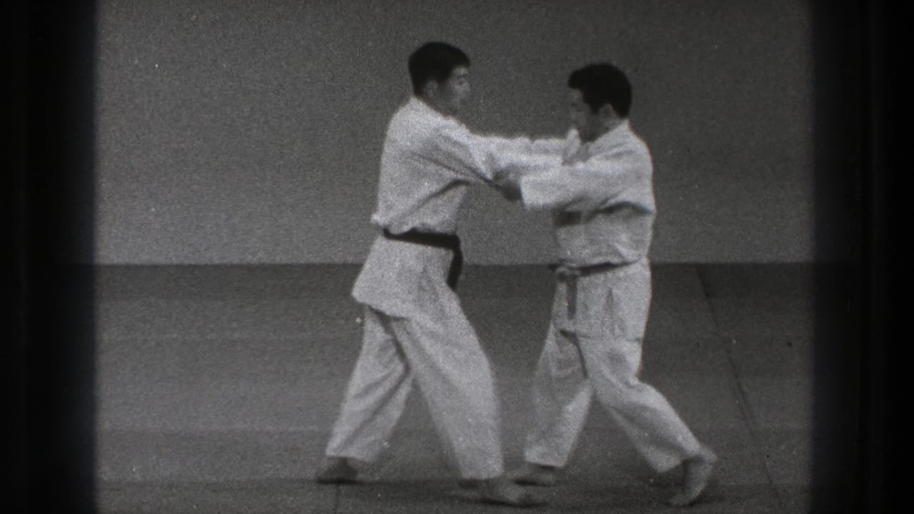 In the black-and-white home-movie frame, the two men maintain arm contact while shifting stance in a karate practice sequence.