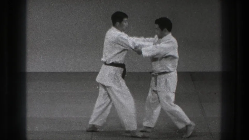 In the black-and-white home-movie frame, the two men maintain arm contact while shifting stance in a karate practice sequence.