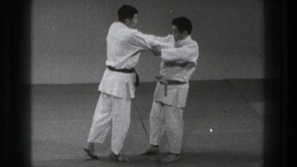 In a Tokyo dojo setting, two adult practitioners in white gi face each other on tatami mats at the start of a throw sequence.