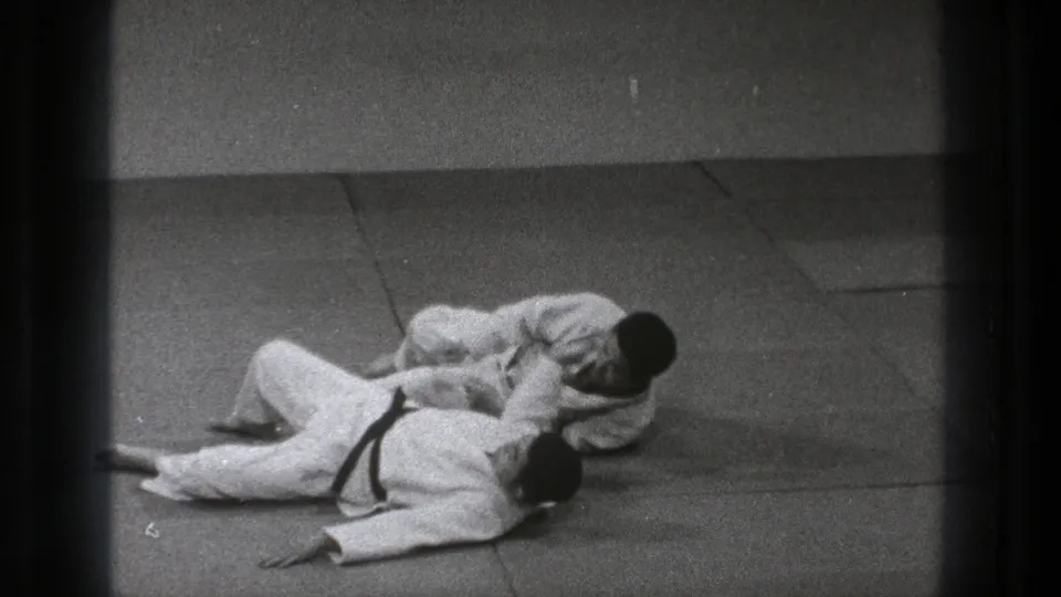 Both practitioners remain low on the tatami in a follow-through position after the throw, with one controlling the other's upper body.