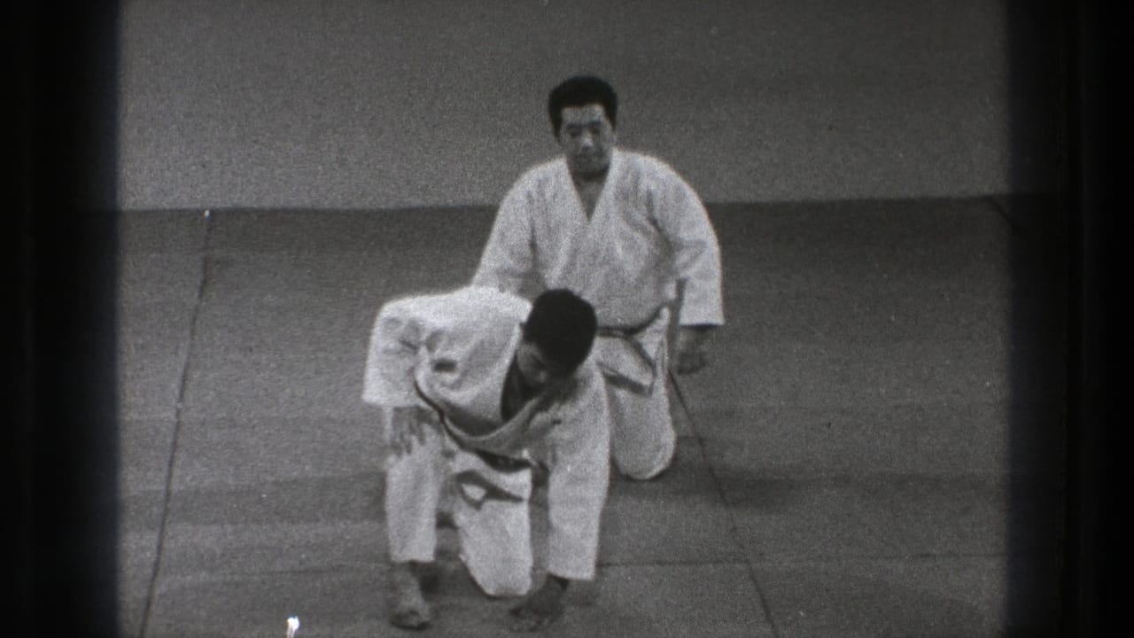 Two men in judo uniforms on a tatami mat, with one kneeling behind another who is rising.
