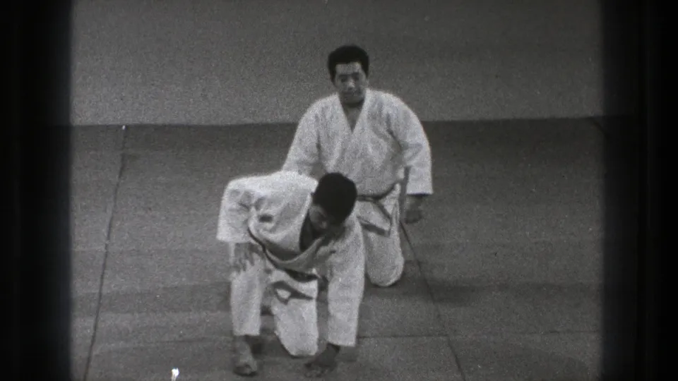In 1971 Tokyo footage, two adult men in white judo gis practice on a tatami mat as one rises from a kneeling position.
