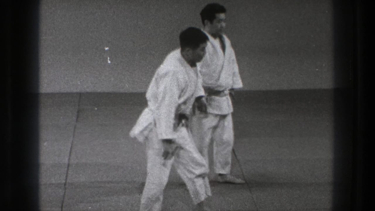 In the Tokyo judo practice scene, two adult men in white gis stand on the mat after a training movement or reset.
