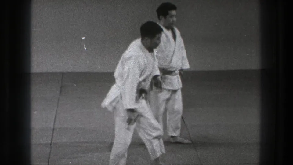 In the Tokyo judo practice scene, two adult men in white gis stand on the mat after a training movement or reset.