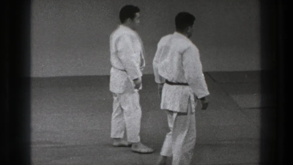 Two adult men in white judo gis stand on the tatami in a Tokyo training space, appearing to reset for another movement.