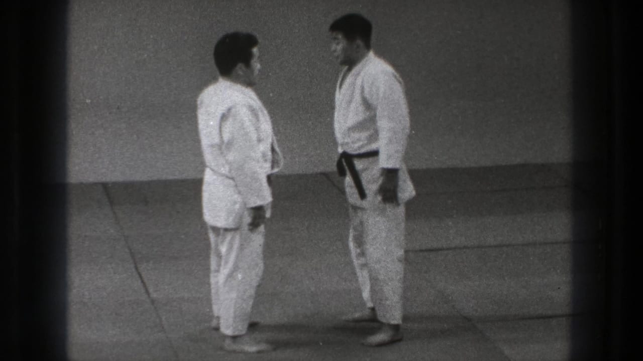 Two judo practitioners stand close together on the mat, one wearing a dark belt.
