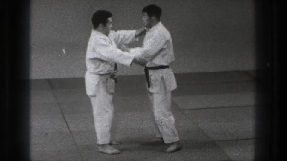 In the Tokyo dojo scene, the practitioners hold each other's gi as the demonstration moves into a controlled contact position.