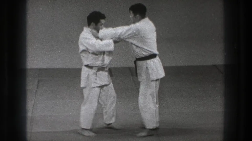 In 1971 Tokyo footage, two barefoot martial artists in white uniforms face each other and grip sleeves on a dojo mat.