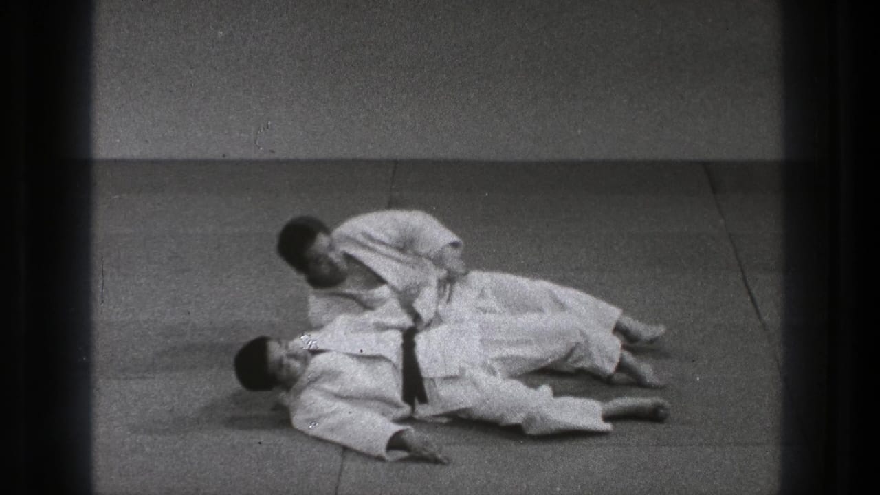 In the closing frame, one Tokyo judo practitioner kneels beside or over the other after the throw on a plain mat.