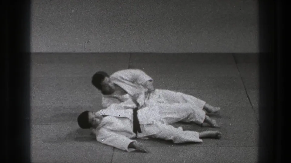 In the closing frame, one Tokyo judo practitioner kneels beside or over the other after the throw on a plain mat.