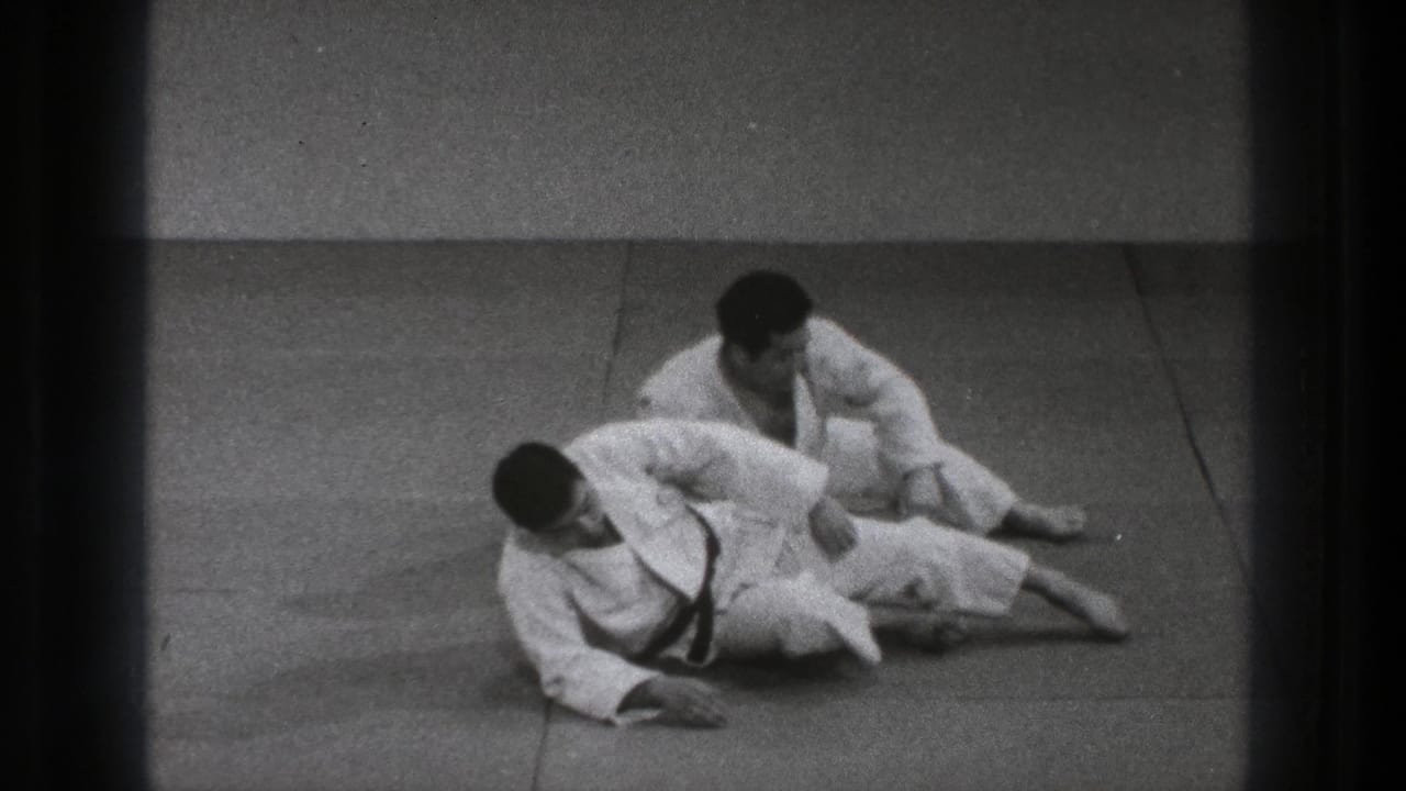 Two men in white gi grapple on a tatami mat, one partly pinned on the floor.