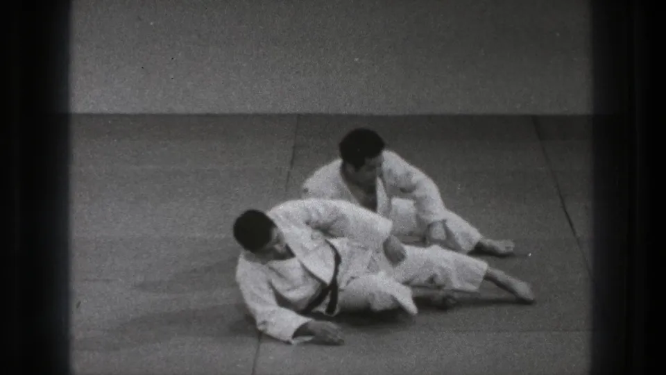 In 1971 Tokyo home-movie footage, two adult men in white gi practice a ground technique on a tatami mat.