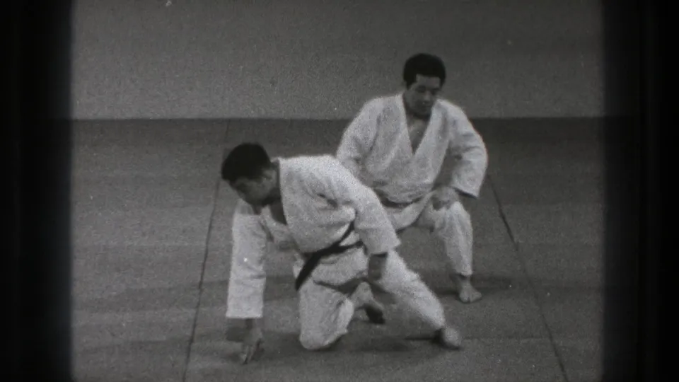 A martial arts practitioner rises to one knee as his partner remains seated on the tatami in 1971 Tokyo footage.