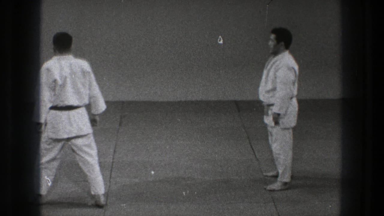 Two martial arts practitioners face one another across a plain tatami training area in the closing moment of the clip.