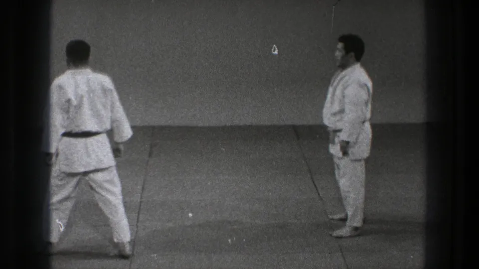 Two martial arts practitioners face one another across a plain tatami training area in the closing moment of the clip.