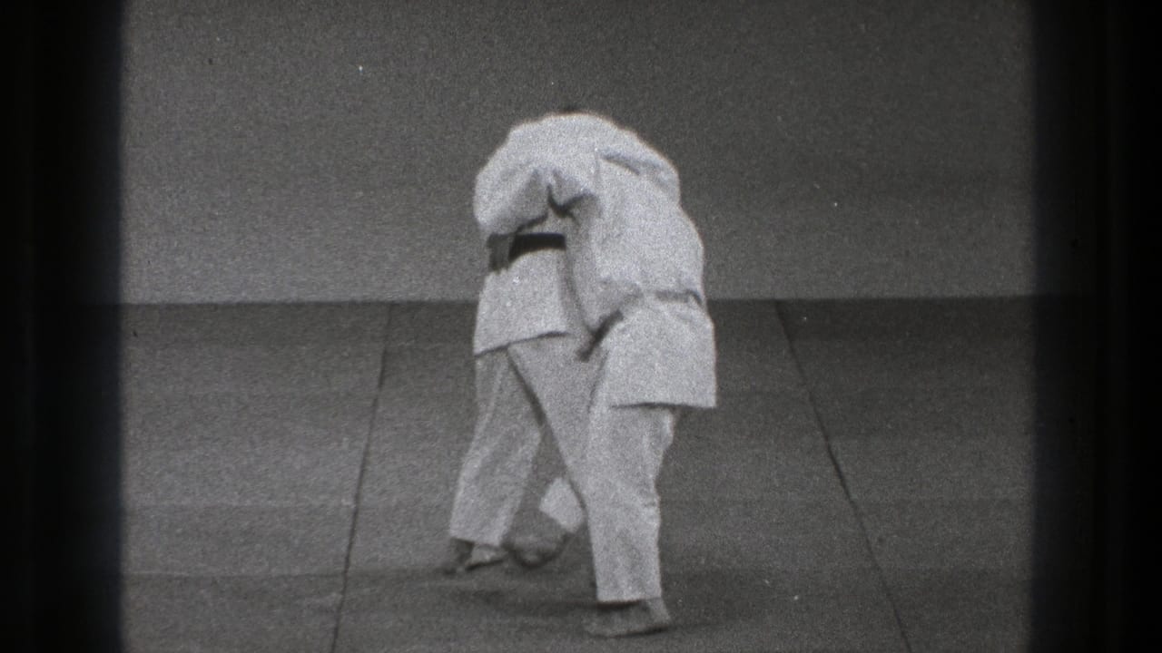 Two men in white gi grapple on a mat near a plain wall.
