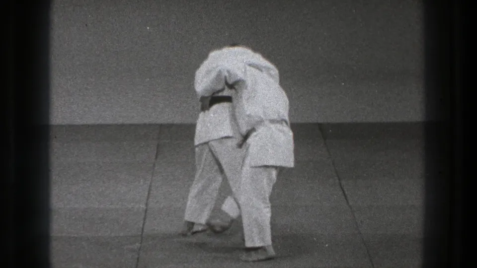 In 1971 Tokyo footage, two men in white gi appear to practice a martial arts hold on a padded mat.