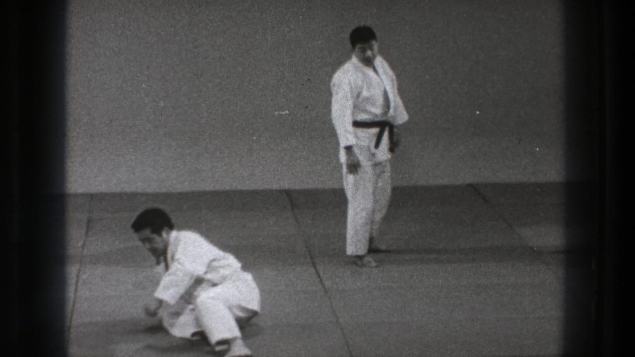 One man sits on the padded floor while another in a dark belt stands nearby during martial arts practice in Tokyo.