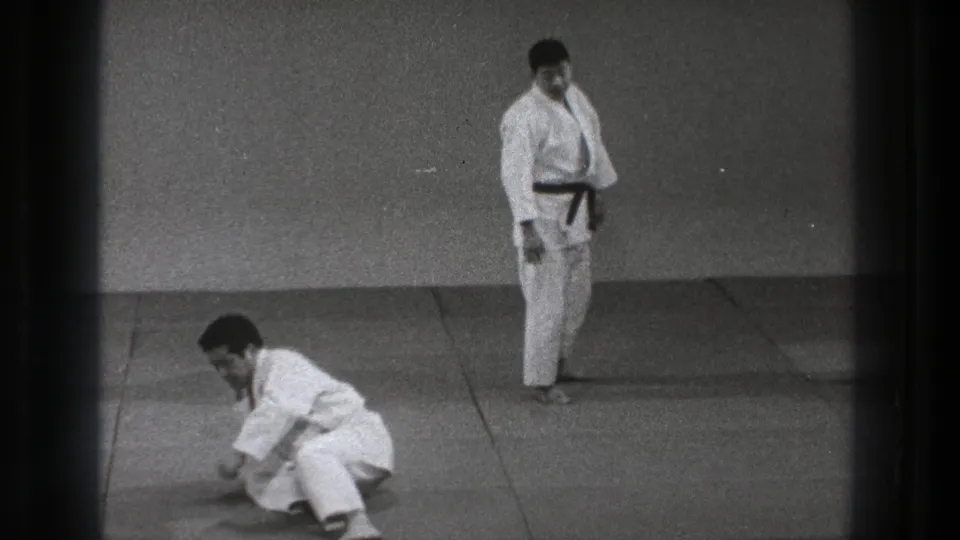 One man sits on the padded floor while another in a dark belt stands nearby during martial arts practice in Tokyo.