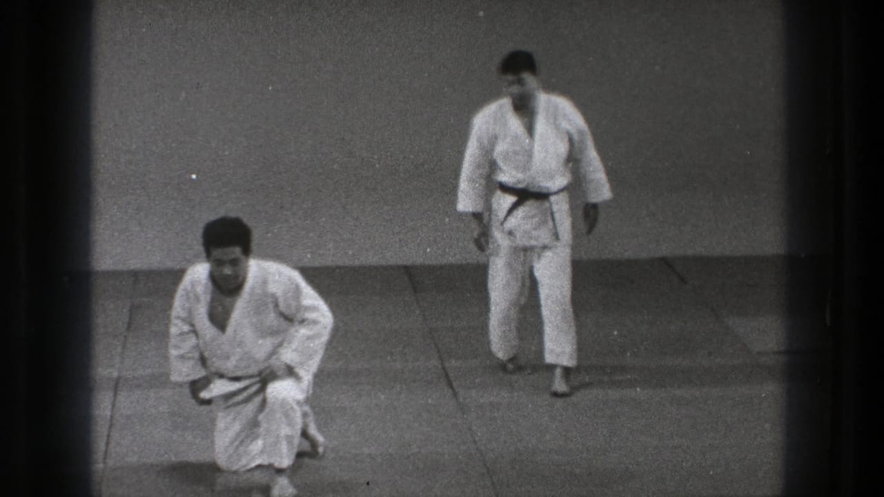 Two men in white gi on a matted dojo floor, one kneeling while the other stands.