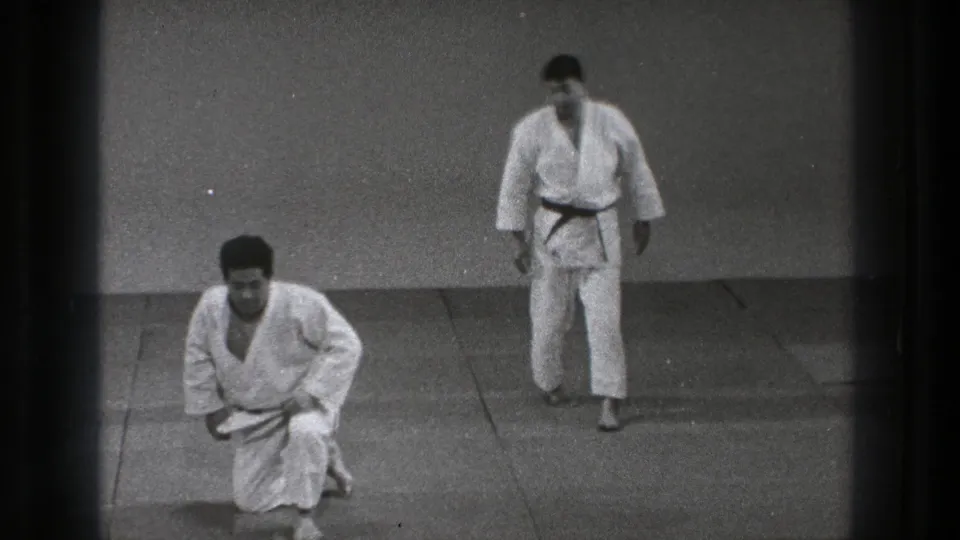 1971 Tokyo dojo footage showing one man kneeling in a white gi as another adult man in a gi stands nearby on the mat.