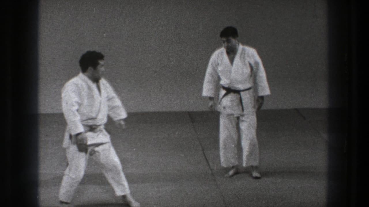 In the 1971 Tokyo karate scene, one practitioner moves into a low stance as the black-belted man observes from across the mat.