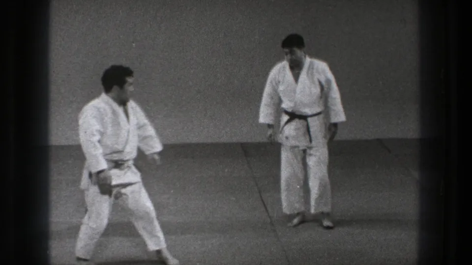 In the 1971 Tokyo karate scene, one practitioner moves into a low stance as the black-belted man observes from across the mat.
