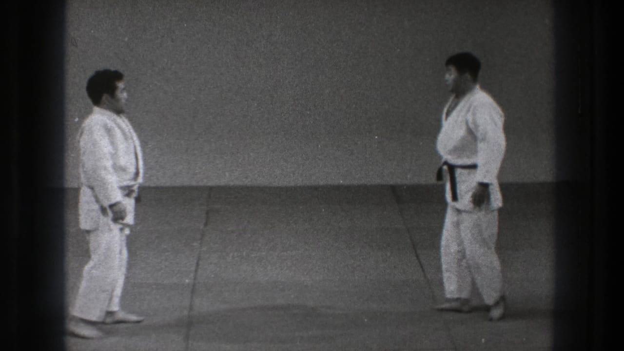 Two adult men in white gi stand facing one another in a Tokyo dojo, with the black belt visible on the right-hand figure.