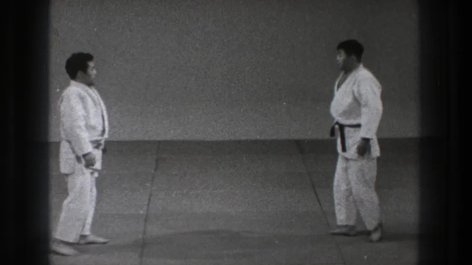 Two adult men in white gi stand facing one another in a Tokyo dojo, with the black belt visible on the right-hand figure.
