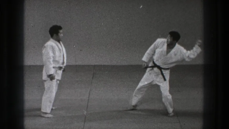 In the 1971 Tokyo clip, two adult men in white gi stand on a plain mat as one black-belted practitioner prepares a movement.