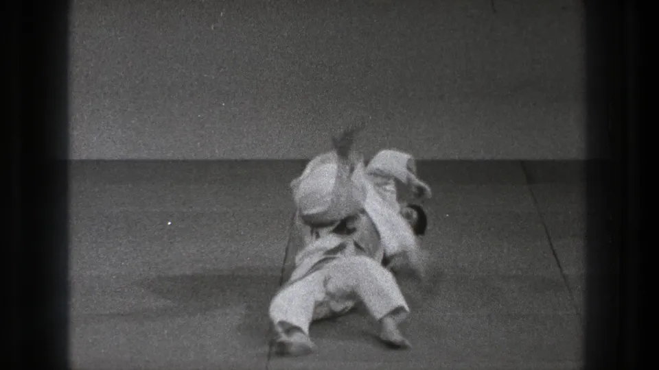 During the 1971 Tokyo practice, one man in a white gi is taken down on the mat while the other remains over him in motion.