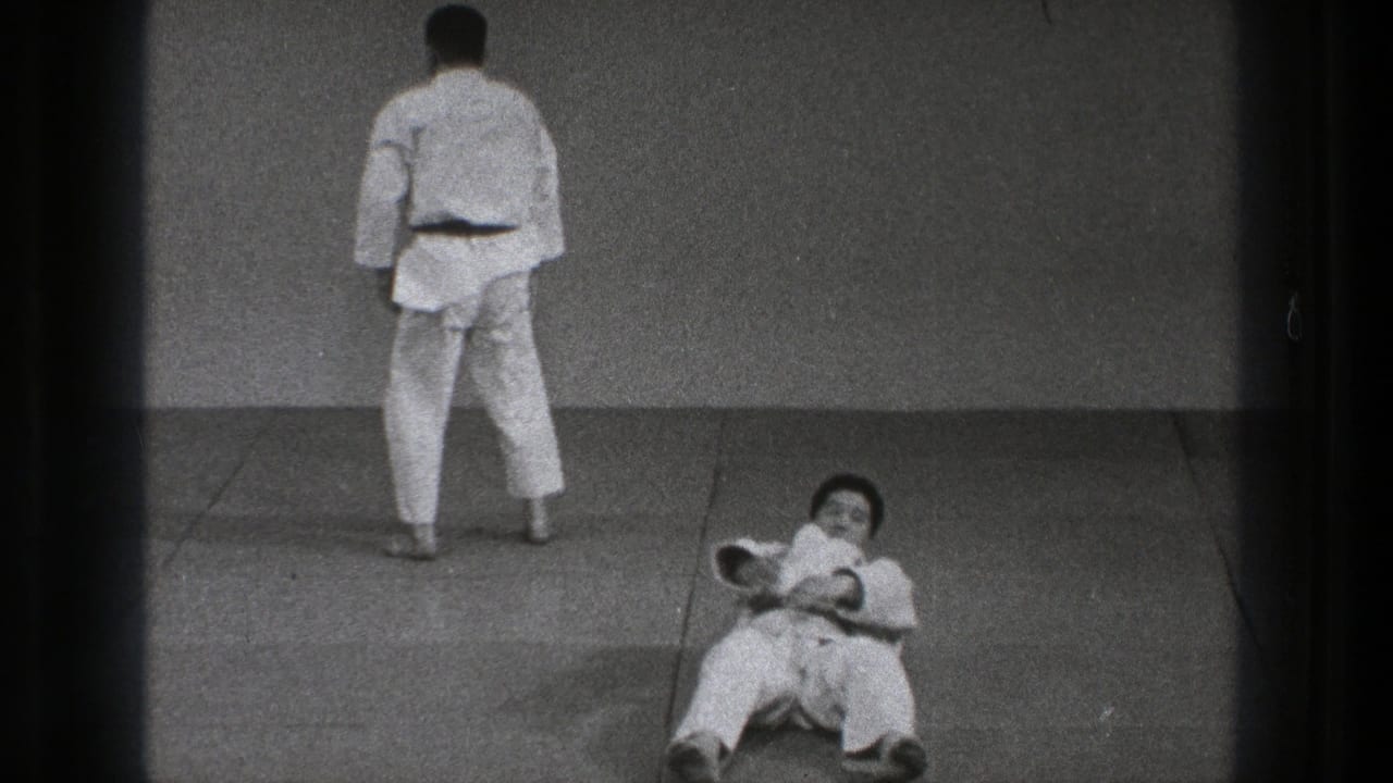 Two men in white gi on mats, one standing with his back turned and one seated after a fall.