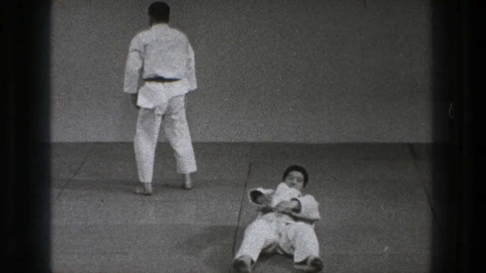 1971 Tokyo karate practice frame showing one adult in a white gi standing near the wall while another sits on the mat after falling.