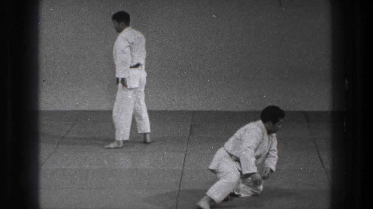 1971 Tokyo karate practice frame with one adult crouching or kneeling after a fall and another white-gi practitioner standing on the mat.