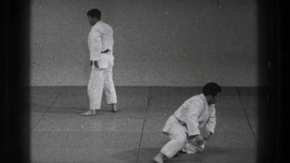 1971 Tokyo karate practice frame with one adult crouching or kneeling after a fall and another white-gi practitioner standing on the mat.