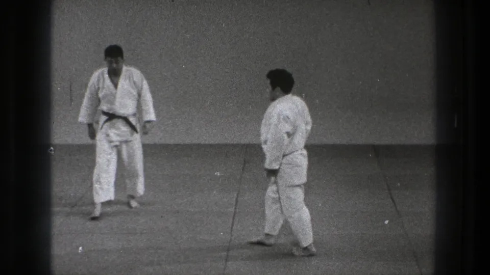 1971 Tokyo karate practice frame with two adult practitioners in white gi standing face to face on a simple indoor mat.