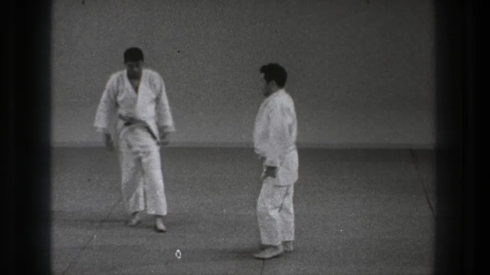 Two adult karate practitioners face one another in a plain Tokyo dojo setting, one wearing a dark belt, 1971 metadata supported.