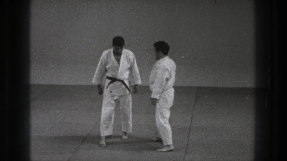 In a 1971 Tokyo karate practice scene, the dark-belt practitioner steps toward a partner in a white gi on a simple mat.