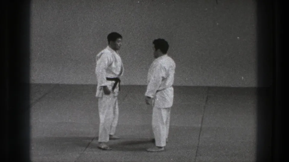 Two adult karate practitioners pause face to face in a plain dojo-like room, their belts and white gi visible in the 8mm frame.