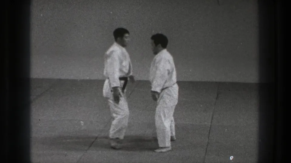 Two adult practitioners in white gi face each other on a mat in the Tokyo 1971 karate footage, with visible film grain and gate edges.