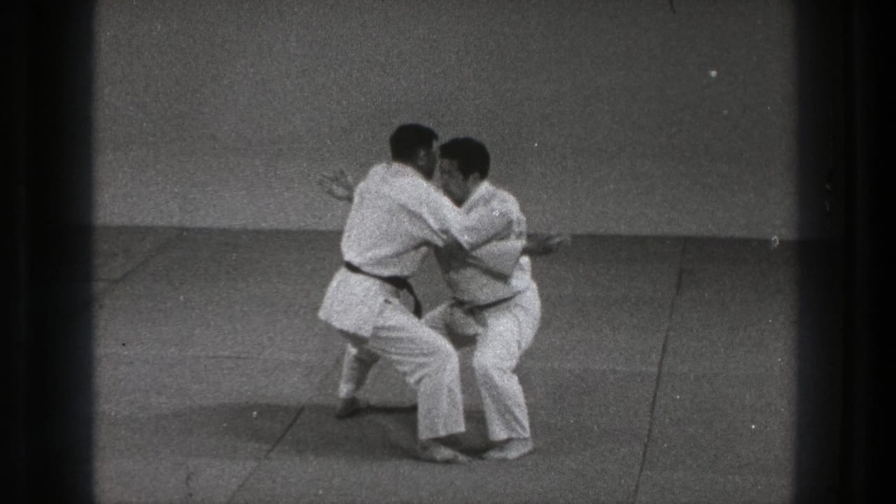 Two men in white gi and dark belts face each other in a low grappling stance on a mat.