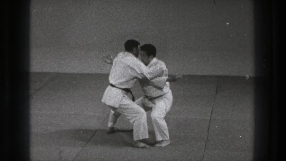 In Tokyo, 1971, two adult karate practitioners in white gi and dark belts begin a close grab on a plain mat.