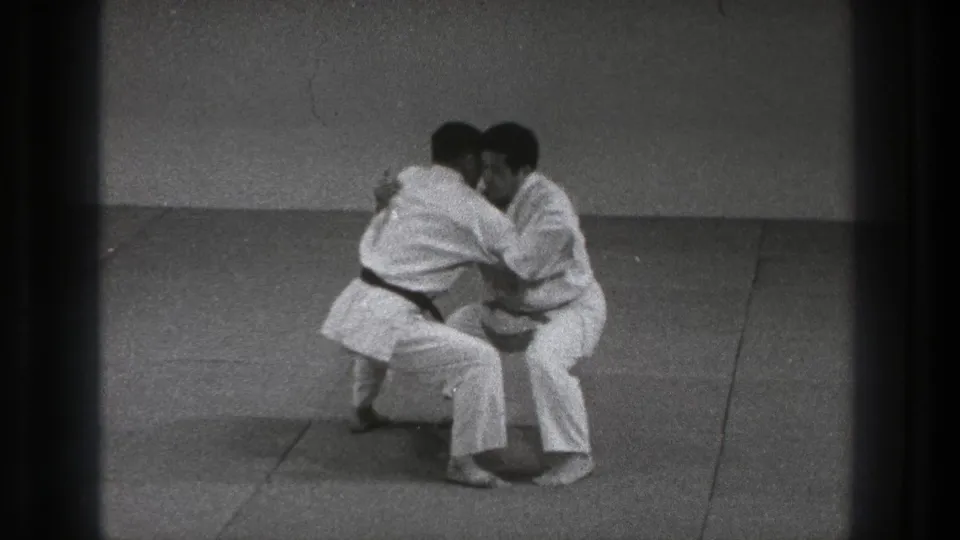 Two adult karate practitioners in Tokyo, 1971, maintain a tight grappling hold while lowering their stance.