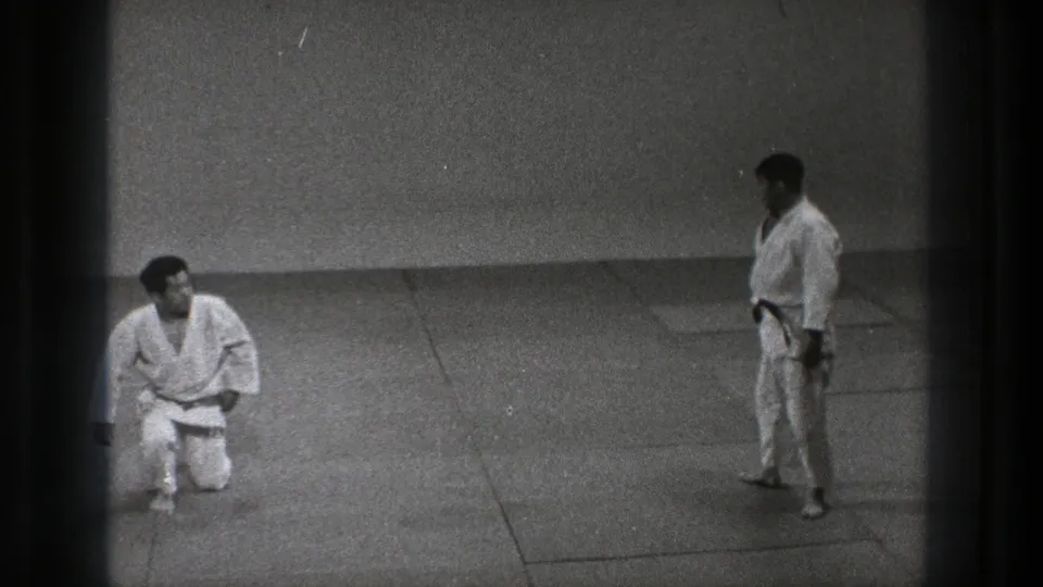 Tokyo 1971 8mm frame with one martial artist kneeling and another standing across the dojo mat.