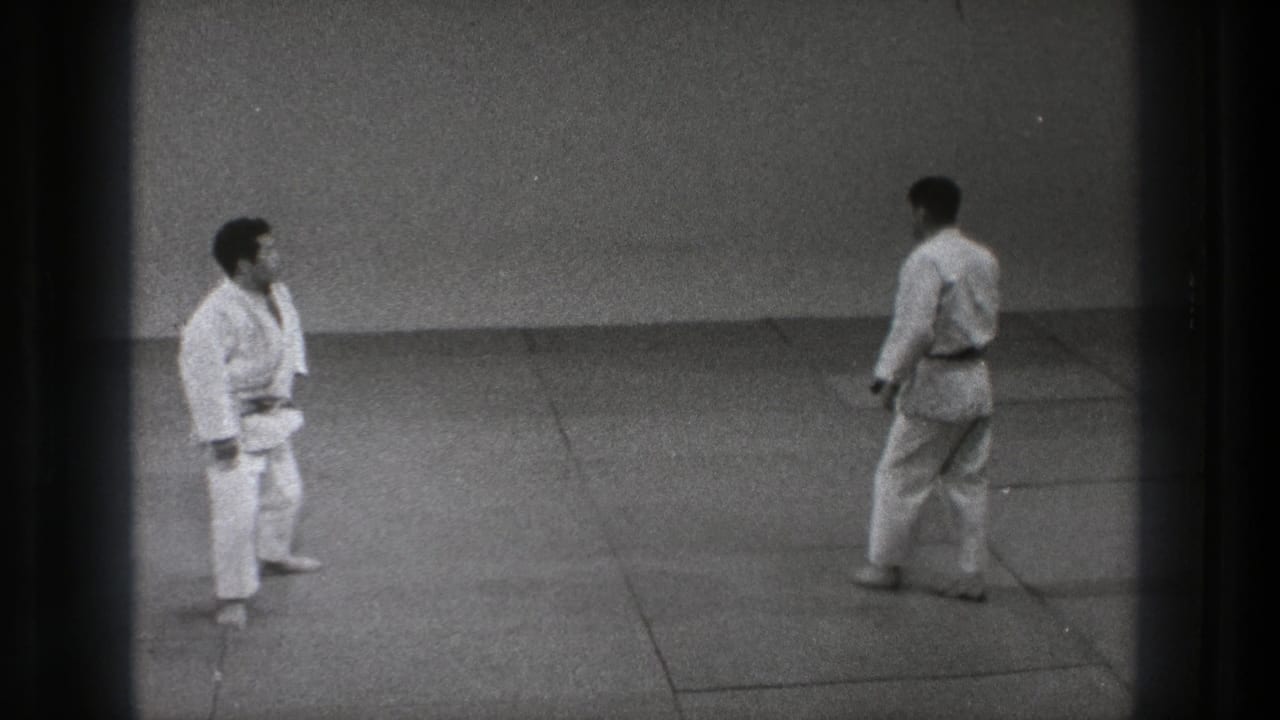 Two martial artists in white gi stand apart on mats inside a plain dojo space.